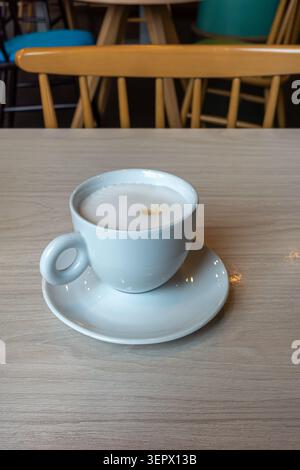Eine Tasse Kaffee steht auf einem Tisch in einem Café. Der Becher ist weiß und hat ein kleines Design. Es gibt Holzstühle und einen farbenfrohen Hintergrund. Es ist mehr Stockfoto