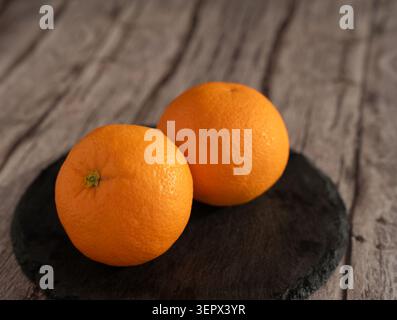Zwei reife Orangen auf einer runden schwarzen Schieferplatte auf einer verwitterten Holzplatte, warmes natürliches Licht, das die Struktur der gekerbten Rinde und vibra hervorhebt Stockfoto