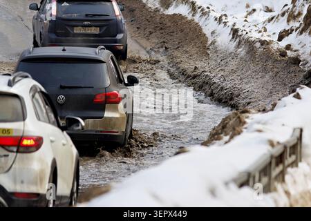 Autos, die während des Auftauens im Frühling eine tiefe, schlammige Pfütze auf einer schlammigen Straße fahren. Schneefälle säumen die unordentlichen Schultern und reflektieren anspruchsvolle Fahrbedingungen Stockfoto