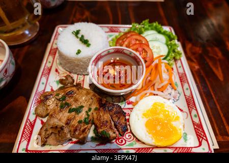 Com tam vietnamesischer Bruchreis mit gegrilltem Schweinekotelett, Spiegelei, eingelegtem Gemüse und Fischsauce, serviert auf traditioneller Keramikplatte Stockfoto