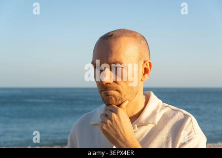 Nachdenklicher Mann nach Haartransplantation, der in Gedanken am Strand von Portugal wegschaut Stockfoto