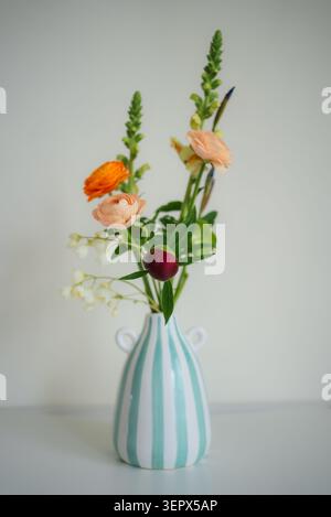 Lebhaftes Stillleben mit einem frischen Frühlingsblumenarrangement mit verschiedenen Blumen und Pfingstrose in einer weiß-türkis gestreiften Vase, die drinnen eine erfrischende Atmosphäre schafft Stockfoto