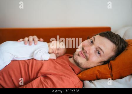 Vater liegt auf dem Rücken, lächelt in die Kamera, während sein neugeborener Sohn auf der Brust schläft und die besondere Verbindung zwischen Eltern und Kind zeigt Stockfoto