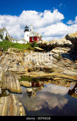 Pemaquid Point Lighthouse und sein rotes Glockenhaus ragen über gestreiften Felsen, die sich in einem stillen Gezeitenbecken unter einem hellblauen Himmel spiegeln. Stockfoto