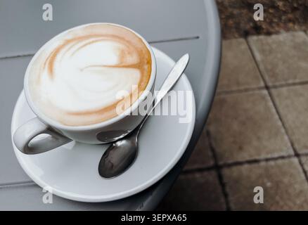 Frischer Cappuccino, serviert in einer weißen Tasse mit Untertasse an einem Außentisch eines Cafés. Kaffeepause, Entspannung und Stadtleben-Konzept. Stockfoto