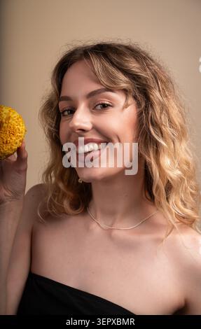 Frau, die hell lächelt, einen lebendigen Zitronenkrapfen hält und Freude und süßen Genuss mit welligen Haaren verkörpert Stockfoto