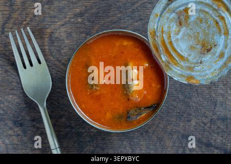 Öffnen Sie die Dose Fisch in Tomatensoße mit Gabel auf Holztisch. Draufsicht auf geöffnete Sardinen oder Sprossen in roter Tomatensauce in einer Dose auf einem Stockfoto