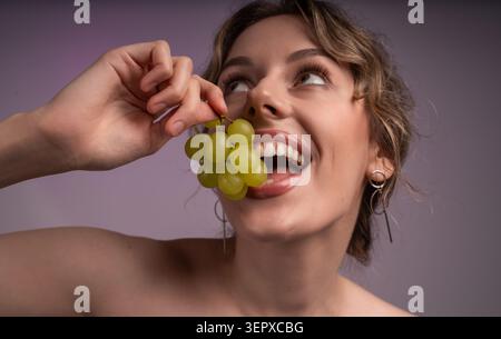 Glückliche Frau, die reife grüne Trauben isst, zeigt gesunde Gewohnheiten und natürliche Schönheit mit einem fröhlichen Ausdruck Stockfoto
