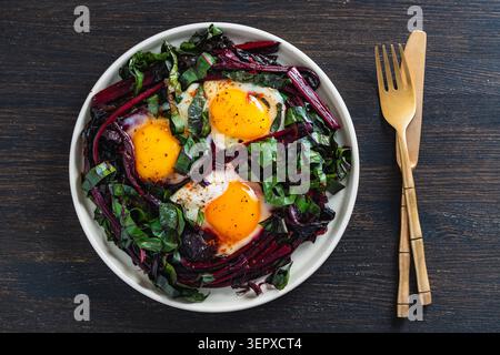 Ein lebhaftes, hochwinkliges Foto von drei sonnigen Eiern, die mit einer großzügigen Hilfe von roten Rübenstämmen und dunkelgrünen Blättern in weiß gebraten werden Stockfoto