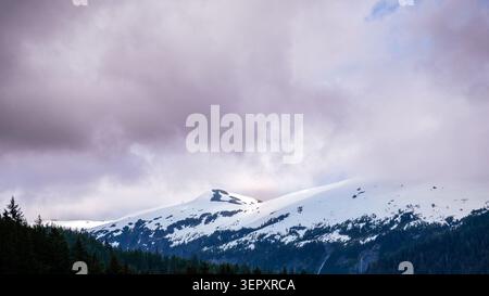 Schnee auf Berggipfeln mit niedrigen Wolken und schattigem Wald Stockfoto