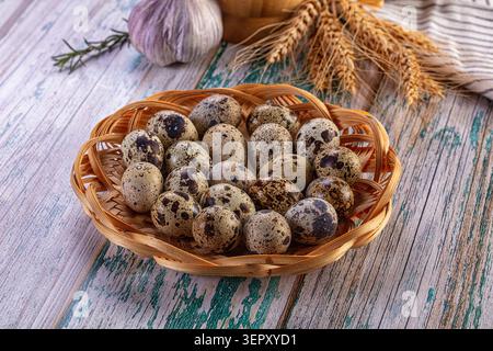 Frisch gesprenkelte Wachteleier in rundem Korb auf bemalter Holzoberfläche mit getrockneten Weizenstielen und Knoblauchzwiebeln in der Nähe von natürlichem li angeordnet Stockfoto