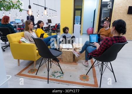 Eine Gruppe junger Profis versammelte sich in einem farbenfrohen Loungebereich. Zwei Frauen sitzen mit einem Tablet auf einem hellgelben Sofa, während Kollegen auf einer Sitzbank sitzen Stockfoto