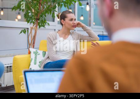 Eine mittlere Aufnahme, die sich auf eine Frau in einer beigen Strickjacke konzentriert, die auf einem gelben Bürosofa sitzt. Sie hört einem Kollegen aufmerksam zu (verschwimmt im Voraus Stockfoto