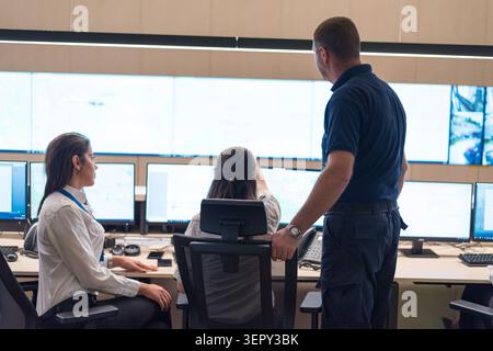 Ein männlicher Sicherheitsvorgesetzter steht hinter zwei weiblichen Operatoren und zeigt auf Daten und Karten auf einer großen Anzeigefand in einem modernen Befehls- und Kontrollzentrum Stockfoto