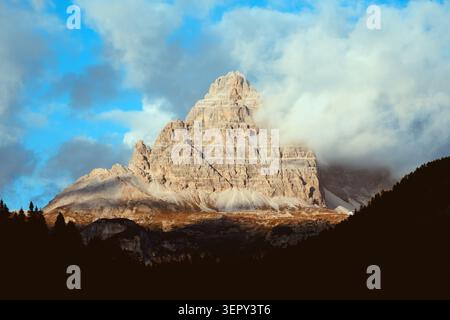 Die sonnenbeschienene Tre Cime di Lavaredo erhebt sich über dunkel bewaldete Hänge, während Wolken um den zerklüfteten Kalksteingipfel driften. Stimmungsvolle Berglandschaft in den Dolomiten Stockfoto