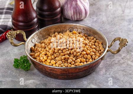 Rohe Sojabohnen in einer alten Kupferschale mit Griffen, umgeben von Knoblauch, Pfeffermühlen, Chili und Petersilie auf einer grauen Steinoberfläche Stockfoto