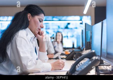 Profilansicht einer jungen Frau in Sicherheitsuniform mit Hilfe einer Computermaus zur Durchsuchung von Videoarchiven in einem dunklen Überwachungsraum. Stockfoto