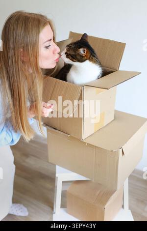 Lächelnde Tierbesitzerin interagiert mit ihrer Katze zwischen Umzugskartons in einer hellen Wohnung, was Umsiedlung und Neuanfänge symbolisiert. Stockfoto