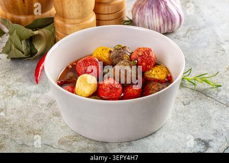 Verschiedene rote gelbe und schwarze Oliven, in Öl mariniert, mit frischen Rosmarin-Lorbeerblättern und hölzernen Mahlwerken auf Steinoberfläche Stockfoto