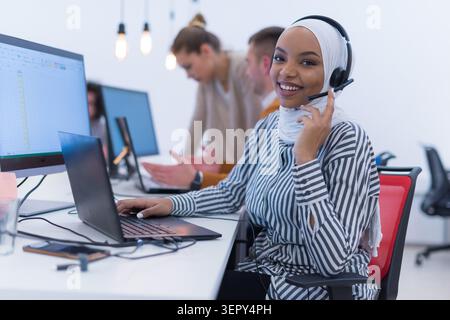 Eine junge Frau, die einen Hijab und ein Headset trägt, lächelt warm, während sie an einem Laptop in einem geschäftigen, modernen Großraumbüro arbeitet. Stockfoto
