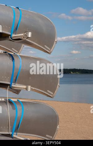 Eine vertikale Nahaufnahme von drei silbernen Metallkanus, die auf einem Regal an einem Strand am See unter blauem Himmel gestapelt sind, mit viel Kopierraum für Text. Stockfoto