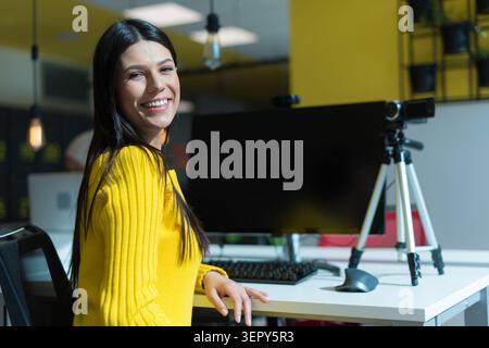 Eine schöne junge hispanische oder kaukasische Frau, die einen hellgelben gerippten Pullover trägt, lächelt breit, während sie an ihrem Schreibtisch sitzt. Ihre Workstation Stockfoto