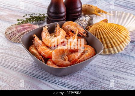 Eine Schüssel mit gekochten Orangengarnelen steht auf einem rustikalen Holztisch, umgeben von Muscheln, Krabbenkrallen, frischen Pfeffermühlen aus Thymian-Holz. Die Szene erinnert an frisches Meer Stockfoto