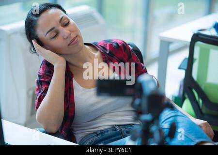 Eine junge Frau, die ihren Kopf auf die Hand lehnt, liegt ruhig mit geschlossenen Augen an ihrem Arbeitsplatz. Sie trägt ein lässiges rotes kariertes Hemd über einem weißen T-Shirt Stockfoto