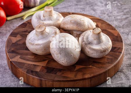 Rohe Champignonpilze auf rundem Holzbrett mit Tomaten und grünen Zwiebeln im Hintergrund. Gesunde vegetarische Zutaten auf rustikalem KI Stockfoto