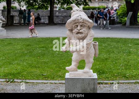 Eine barocke Steinstatue eines schreitenden Zwergs mit Spitzmütze und Bart im Zwerggarten der Mirabellgärten in Salzburg, mit zahlreichen Besuchern im Hintergrund. Der Zwerggarten ist eine der meistfotografierten Attraktionen in den Mirabellgärten in Salzburg, Österreich Stockfoto