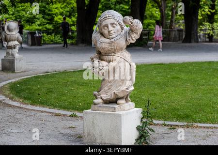 Eine barocke Steinskulptur einer Zwergfigur mit Kopftuch, Obst in beiden Händen und mit Obstkorb vor ihr, im Zwerggarten des Mirabellgartens in Salzburg; weitere Skulpturen des Ensembles sind im Hintergrund zu sehen. Die weiblichen Figuren im Zwergerlgarten sind mit Symbolen der Ernte und Fruchtbarkeit dargestellt und bilden einen Kontrast zu den meist karikierten, männlichen Grotesken. Mirabell Gardens, Salzburg, Österreich Stockfoto