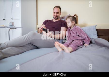 Vater und Tochter entspannen sich im Schlafanzug auf dem Bett, schauen gemeinsam auf das Smartphone Stockfoto