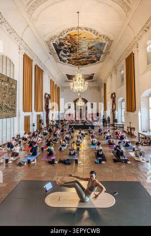 Ein Yogalehrer demonstriert eine Seitenstrecke auf der Bühne, die Teilnehmer im Ballsaal des DomQuartiers Salzburg folgen der Pose. Ein barockes Deckenfresko und Kristallleuchter sind im Hintergrund zu sehen. Veranstaltung: Pop Up Yoga – Mozart Edition. Salzburg, Österreich Stockfoto