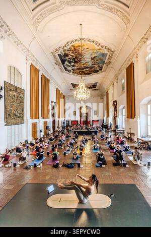 Blick auf den Ballsaal im DomQuartier Salzburg von der Galerie aus: Mehrere Dutzend Teilnehmer sitzen auf Matten; auf dem zentralen Podium führt ein Yogalehrer die Sitzung mit einem Kronleuchter und einem barocken Deckenfresko im Hintergrund. Veranstaltung „Pop Up Yoga – Mozart Edition“. Salzburg, Österreich Stockfoto