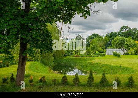 Malerische lettische Landschaft mit einem kleinen Teich, einer grünen Wiese, Bäumen und einem Bauernhof unter dramatischen Sommerwolken Stockfoto