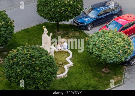 Blick von oben auf eine Steinstatue eines Bischofs mit krozier und Mitra durch ein einfaches Brunnenbecken; zwei junge Frauen sitzen am Rand des Beckens. Die Skulptur steht an einem Grasrand inmitten eines Parkplatzes an der Clemens-Holzmeister-Stiege. Salzburg, Österreich Stockfoto