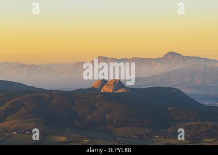 Blick aus der Vogelperspektive auf die zerklüftete Bergkette und Granitgipfel während der goldenen Stunde über das üppige Tal und die Weinberge in Stellenbosch, Western Cape, Süd Stockfoto