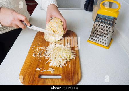 Nahaufnahme einer Frau, die frischen Weißkohl mit einem Messer auf einem Holzbrett neben einer gelben Metallreibe in der Küche schnippelt Stockfoto