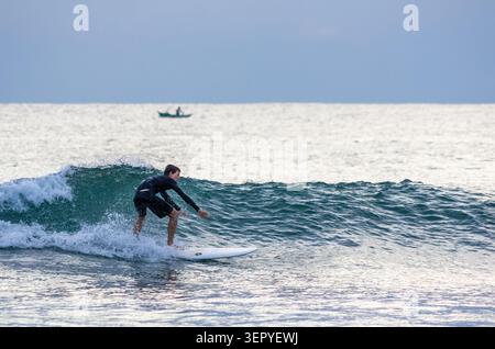 Mirissa, Sri Lanka - 20. Februar 2023: Ein Surfer in einem schwarzen Neoprenanzug reitet am Mirissa Beach auf einer Welle, wobei ein kleines Fischerboot auf dem ruhigen Meer sichtbar ist Stockfoto