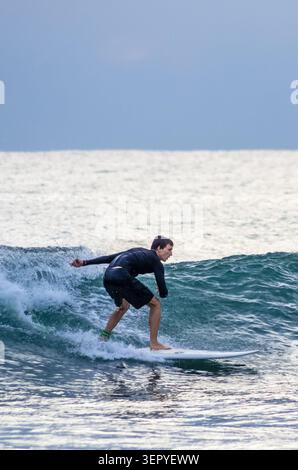 Mirissa, Sri Lanka - 20. Februar 2023: Ein Surfer in einem schwarzen Neoprenanzug reitet auf einem weißen Surfbrett am Mirissa Beach auf einer türkisfarbenen Meereswelle Stockfoto
