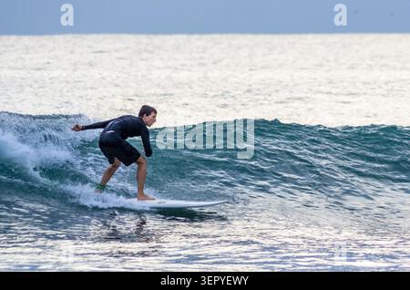 Mirissa, Sri Lanka - 20. Februar 2023: Ein Surfer in einem schwarzen Neoprenanzug reitet auf einem weißen Surfbrett am Mirissa Beach auf einer türkisfarbenen Meereswelle Stockfoto
