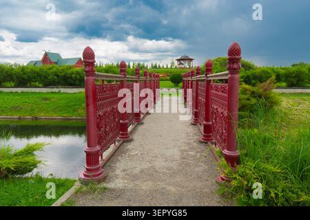 Elegante rote Metallbrücke über einen Bach in einem grünen Garten. Im Hintergrund befindet sich ein Landschaftspark mit Pavillon, Rasen und dramatischem bewölktem Himmel Stockfoto