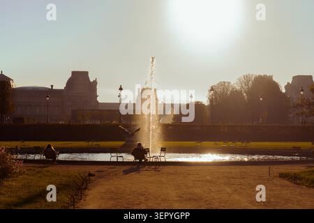 Springbrunnenstrahl am Jardin des Tuileries in Paris während eines trüben goldenen Sonnenuntergangs, mit dem Louvre-Museum im Hintergrund. Die Leute entspannen sich. Stockfoto