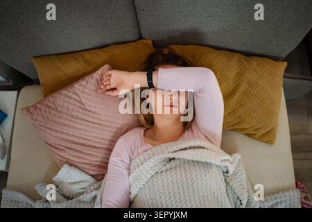 Junge Erwachsene Frau, die ihre Augen mit einem Arm bedeckt, während sie auf einem Sofa ruht und sich unwohl fühlt Stockfoto