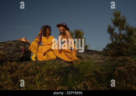 Zwei Frauen in gelben Kleidern sitzen bei Sonnenuntergang auf einem felsigen Hügel Stockfoto