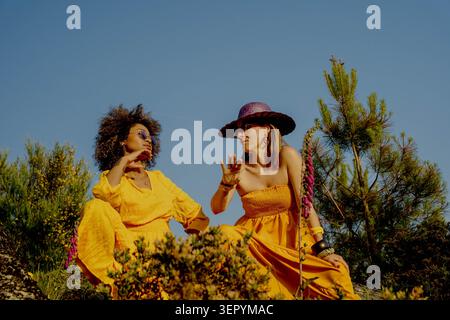 Zwei Frauen in gelben Kleidern plaudern draußen unter Kiefern Stockfoto