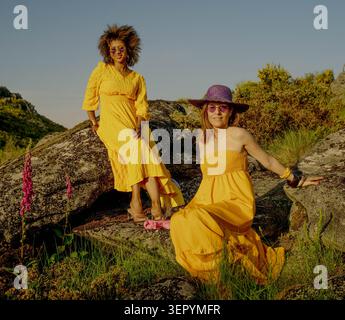 Zwei Frauen in gelben Kleidern posieren auf sonnendurchfluteten Felsen in einer natürlichen Landschaft Stockfoto