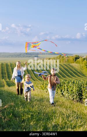 Glückliche Familie, eine Mutter und zwei Söhne, die auf einem grünen Feld laufen, Spaß haben, einen farbenfrohen Drachen zu fliegen und mit einem Spielzeugflugzeug zu spielen Stockfoto