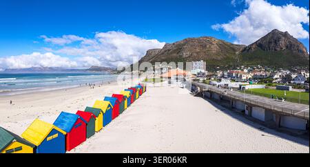Aus der Vogelperspektive sehen Sie die berühmten farbenfrohen Strandhütten am Sandstrand von Muizenberg Beach mit Bergen und einer Promenade in Kapstadt, Westkap, Süd Stockfoto
