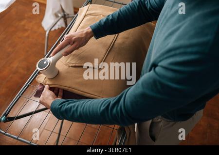 Hochwinkelaufnahme eines Mannes, der Flusen von einem beigen Sweatshirt mit einem modernen elektrischen Stoffrasierer entfernt, während er Hausarbeiten und Kleidungspflege durchführt Stockfoto